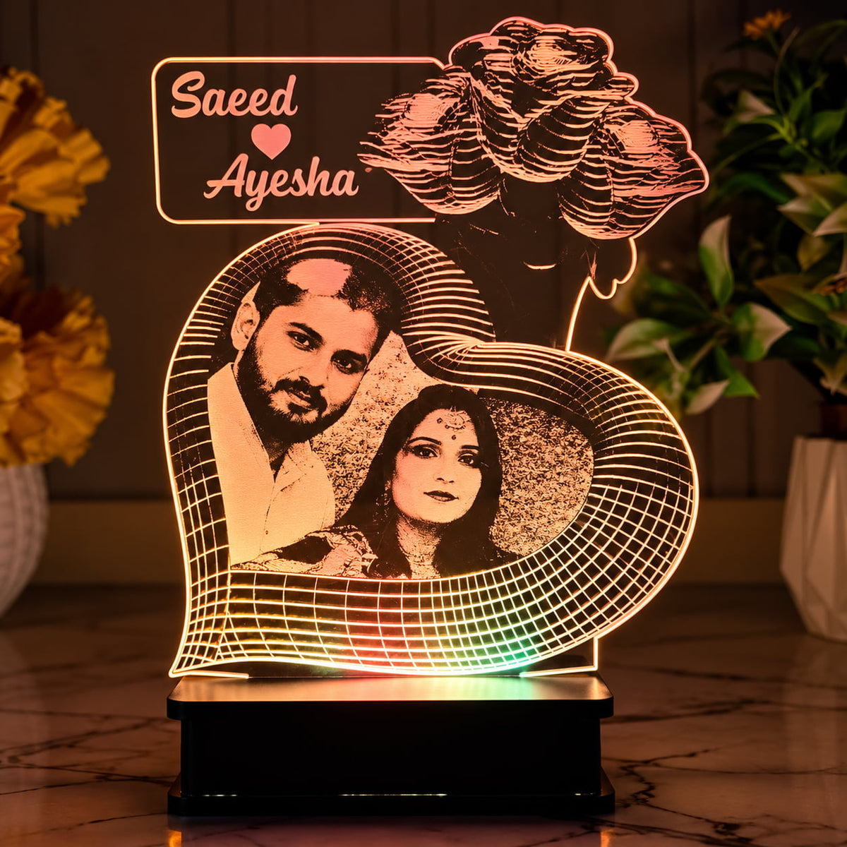A personalized acrylic LED night light shaped like a heart, featuring laser-cut portraits of a couple and the names 'Saeed' and 'Ayesha' with a small heart between them. The light is illuminated with warm pink, orange, and yellow tones, sitting on a black base on a marble-patterned surface with blurred potted plants in the background.