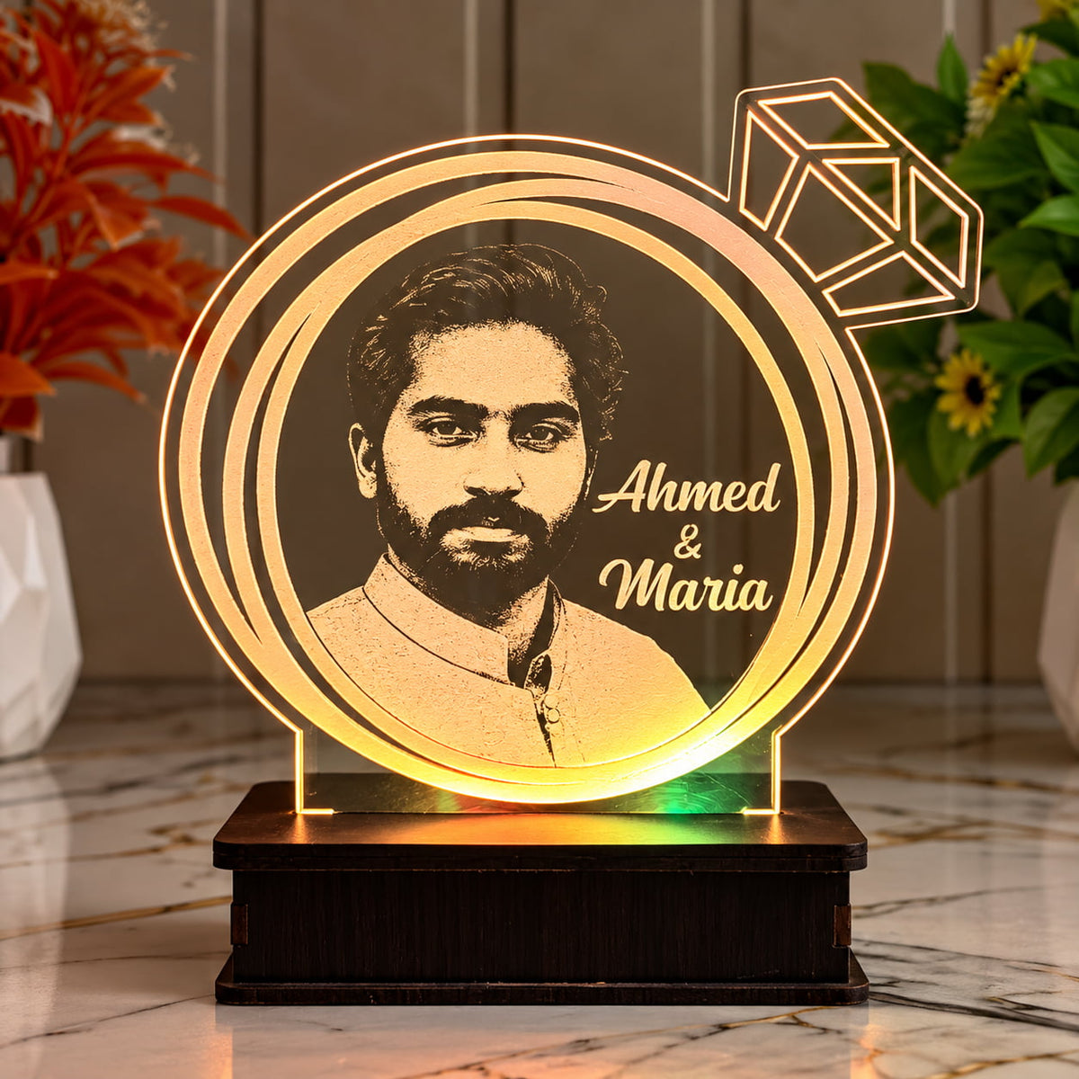 A personalized acrylic LED light shaped like a ring with a portrait of a man and the names 'Ahmed & Maria' engraved, displayed on a dark wooden base on a marble surface with indoor plants in the background.