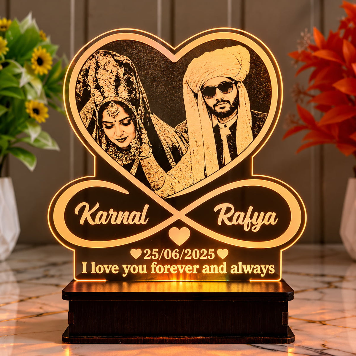 A personalized acrylic wedding plaque on a dark wooden base, featuring a heart-shaped couple portrait, an infinity symbol with the names 'Karnal' and 'Rafya', and the date '25/06/2025' with the message 'I love you forever and always'.