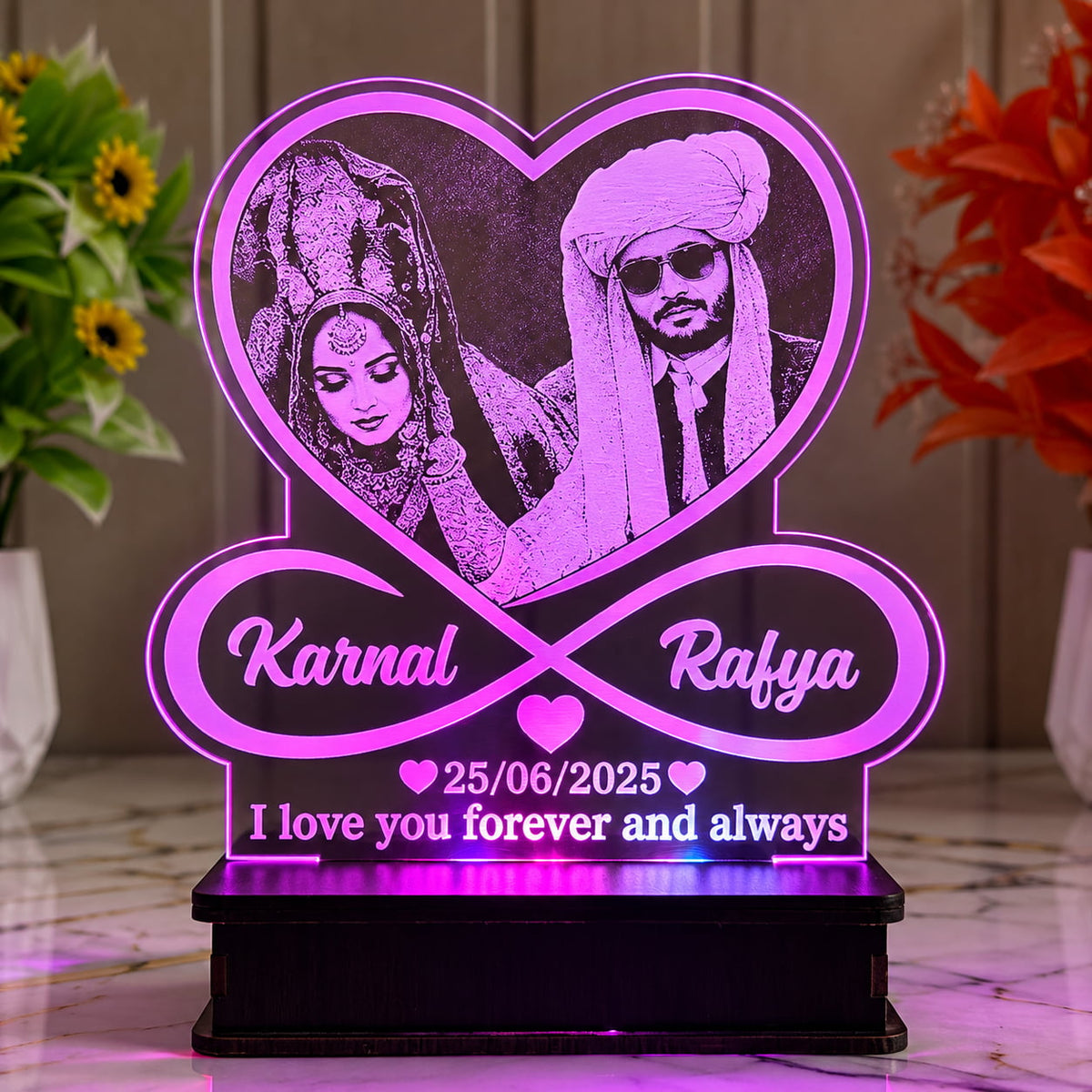 A personalized acrylic LED light table featuring a heart-shaped couple's portrait, an infinity symbol with the names 'Karnal' and 'Rafya', the date '25/06/2025', and the message 'I love you forever and always', illuminated in pink and purple tones on a black base.